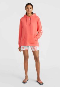 Koraliowy hoodie z miękkiego materiału z kieszenią z przodu, zestawiony z szortami w tie-dye w jasnych kolorach oraz klapkami z różowym kokardkowym detalem.