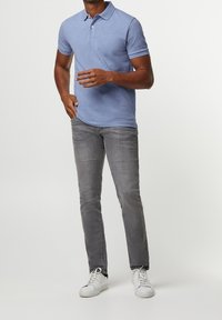 Světle modré polo tričko s krátkým rukávem, šedé slim-fit džíny a bílé tenisky. Model stojí s gestikulací proti jednobarevnému pozadí.