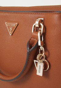 Borsa a tracolla in pelle marrone con una superficie testurizzata, un emblema dorato triangolare, accessori decorativi in oro e un portachiavi con il nome del marchio.