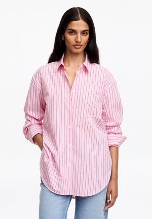 Femme aux longs cheveux foncés porte une chemise rose à boutons avec de fines rayures verticales rouges et un jean bleu clair, debout avec une main dans la poche.