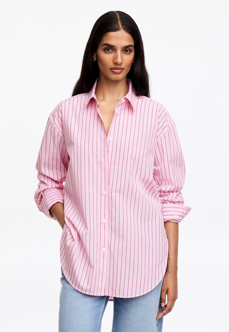 Femme aux longs cheveux foncés porte une chemise rose à boutons avec de fines rayures verticales rouges et un jean bleu clair, debout avec une main dans la poche.