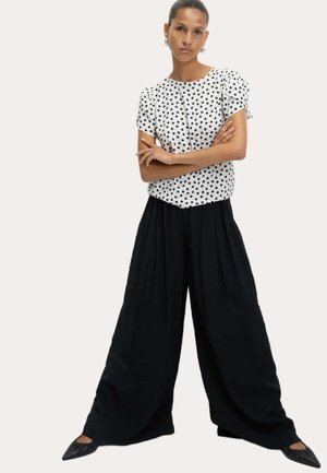 Vit blus med svarta prickar, korta puffärmar, i kombination med lösa svarta plisserade byxor. Svarta spetsiga ballerinaskor fulländar outfiten.