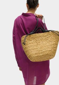 Geflochtene Strohtasche mit weitem, offenen oberen Bereich, schwarzen geflochtenen Griffen und einer natürlichen, tanartigen Farbe. Getragen über einem lila Hemdkleid.