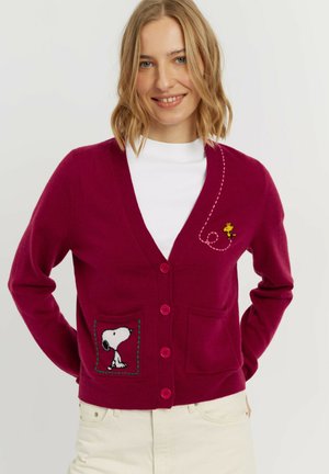 Vrouw die een bordeaux cardigan draagt met Snoopy op de linkerpocket en Woodstock geborduurd nabij de schouder, glimlachend met handen achter de rug.