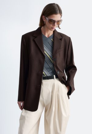 Kvinde iført oversized mørkebrun blazer, stribet grå V-hals sweater, cremefarvede plisserede bukser, sort bælte og store solbriller, poserer med hånden i lommen.