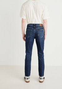 Tumman siniset farkut suoran lahkeen istuvuudella ja nahkainen brändilappu takavyötäröllä, yhdistettynä yksinkertaiseen valkoiseen t-paitaan ja lenkkareihin.