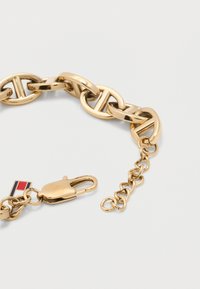 Βραχιόλι αλυσίδας από χρυσό με μεγάλα οβάλ συνδέσμους, κούμπωμα με κρίκο, προέκταση αλυσίδας και μικρή ορθογώνια ετικέτα μάρκας σε κόκκινο, λευκό και μπλε χρώμα.