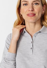 Lichtgrijze polo met een kraag, drie knopen en lange mouwen. Heeft een gestructureerde stof en subtiele stiksels.