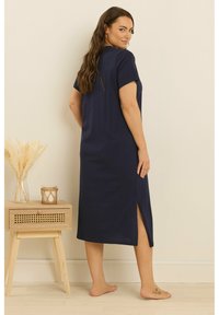 Mörkblå, knälång klänning med korta ärmar, slits på sidan och mjuk textur. Bärs av en person som står bredvid ett träskåp.