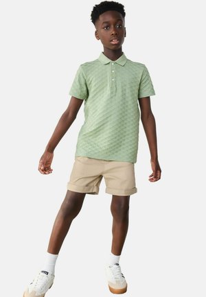 Polohemd in hellgrün mit strukturiertem Muster; Kragen und drei Knöpfe. Kombiniert mit beigen, hochgerollten Shorts und weißen Sneakers.