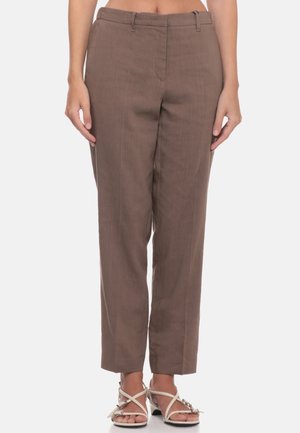 Donna con pantaloni marroni a vita alta e gamba dritta e sandali bianchi con tacco e cinturini, in piedi davanti a uno sfondo semplice.