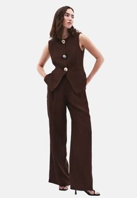 Bruine mouwloze jumpsuit met een aansluitende lijfje, wijde broekspijpen en drie decoratieve knopen. De stof lijkt glad met een lichte glans.