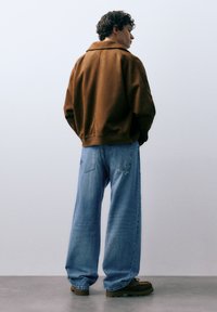 Bruine oversized jas met een kraag, gecombineerd met lichtblauwe jeans met wijde pijpen en bruine schoenen, die een casual en ontspannen pasvorm tonen.