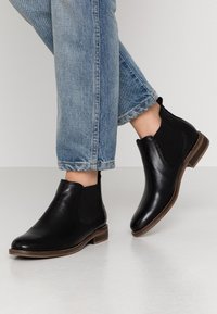 Černé kotníkové boty s texturovanou kůží, elastickými bočními panely a nízkým nasazeným podpatkem, nošené s ohrnutými džíny.