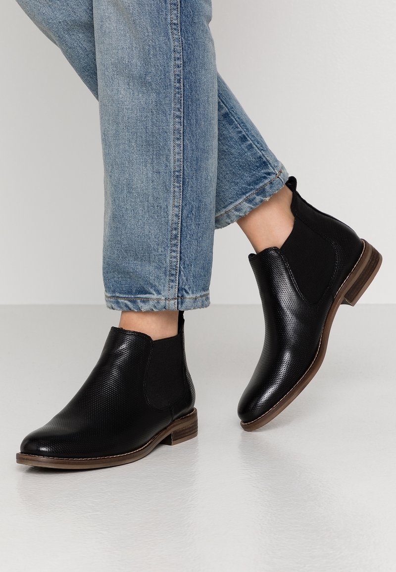 Černé kotníkové boty s texturovanou kůží, elastickými bočními panely a nízkým nasazeným podpatkem, nošené s ohrnutými džíny.