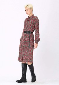 Robe florale rouge à manches longues, avec un bouton-pression devant et un ourlet échancré, cintrée à la taille par une ceinture noire. Portée avec des bottes noires montantes.