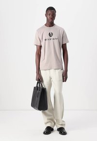 Tan cotton t-shirt with a black "BELSTAFF" logo, paired with off-white wide-leg trousers and a structured black tote bag. Black shoes.