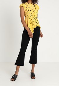Žlutý top s puntíky, s volánovými rukávy a peplum designem, zkombinovaný s černými rozšířenými džíny a černými skluzavkami s kovovým akcentem.