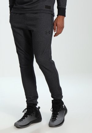 Homme portant un pantalon de jogging gris foncé texturé et des baskets montantes noires et grises, debout sur un fond clair.