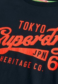 Must mustrit T-särk, millel on erkpunane, praakunud tekst disain "TOKYO Super T JPN 6 HERITAGE CO." Ümmargune kaelus. Tekstuuriga printimise detailid.