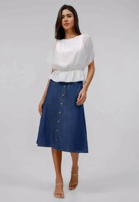 Γυναίκα ντυμένη με λευκή κοντομάνικη μπλούζα με πεπλοειδή μέση και μπλε μεσάτη φούστα με κουμπιά μπροστά, στέκεται με το ένα χέρι στη μέση.