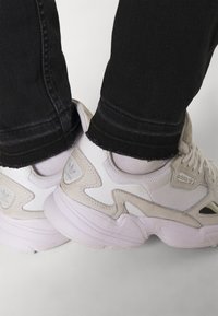 Zapatillas deportivas blancas y beige claro, hechas de materiales de malla y cuero, con acentos texturizados y un logotipo prominente en el costado.
