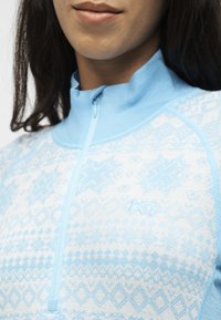 Femme portant un pull zippé bleu clair et blanc à motifs, avec un col montant et un petit logo brodé sur la poitrine.