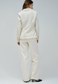 Textured white blazer with frayed hem and gold button accents, paired with cream wide-leg trousers and black pointed heels.