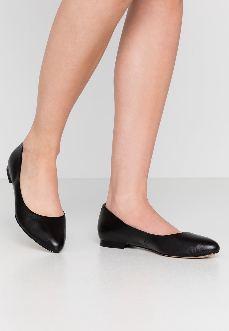 Jambes portant des ballerines simples en cuir noir à talon bas sur un sol blanc avec un fond gris clair.