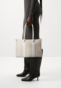 Bolso de mano tejido beige con acentos de cuero blanco y logo grabado. Forma rectangular, doble asa y patrón texturizado. Botas negras hasta la rodilla.
