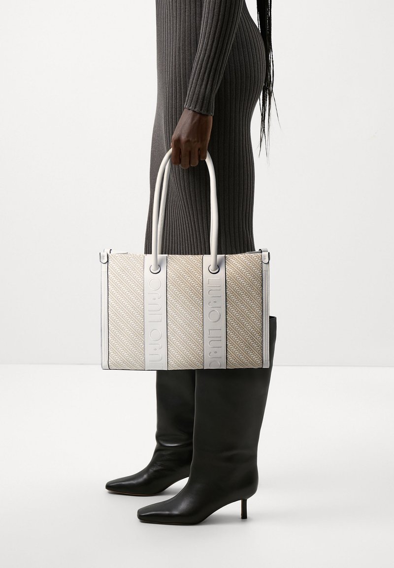 Bolso de mano tejido beige con acentos de cuero blanco y logo grabado. Forma rectangular, doble asa y patrón texturizado. Botas negras hasta la rodilla.