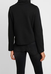 Felpa nera a maniche lunghe con collo a lupetto, design cropped e texture morbida. Presenta un colore uniforme e nessun motivo o accessorio visibile.