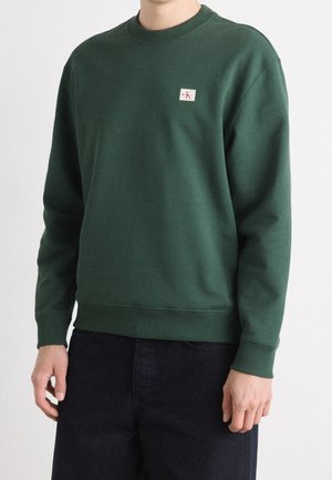 Personne portant un sweat-shirt vert forêt à col rond avec un petit logo Calvin Klein sur la poitrine et un pantalon foncé sur un fond uni.