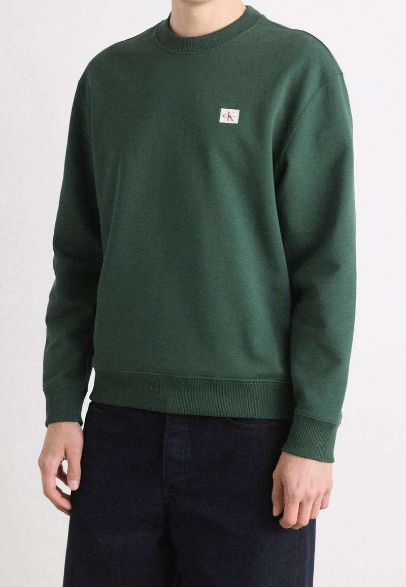 Personne portant un sweat-shirt vert forêt à col rond avec un petit logo Calvin Klein sur la poitrine et un pantalon foncé sur un fond uni.