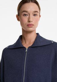 Navy-zip-Jacke aus strukturiertem Strick mit breitem Kragen und weißen Akzentnähten. Verfügt über einen silbernen Reißverschluss und einen lockeren Schnitt.