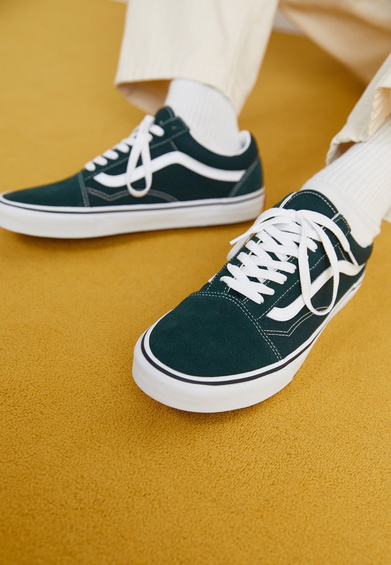 Personne portant des baskets vert foncé et blanches avec des lacets blancs et un pantalon clair sur un sol moquetté jaune moutarde.