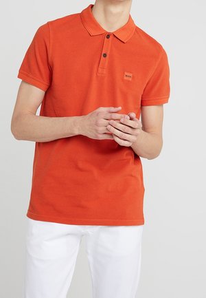 Hombre con una camisa polo de manga corta naranja con botones negros y pantalones blancos, con las manos entrelazadas al frente.