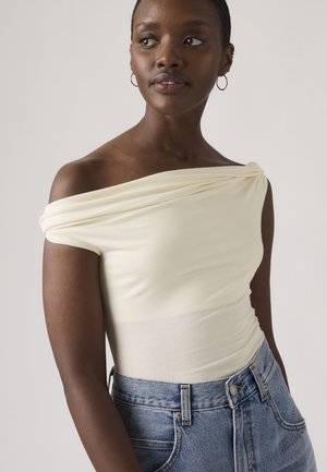 Femme à la peau foncée portant un haut crème à épaules dénudées et un jean bleu, regardant sur le côté sur un fond uni.