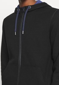 Sweat à capuche noir zippé en tissu doux avec une finition texturée, doté d'une capuche doublée de bleu et de cordons tressés bleus. Poches avant simples.