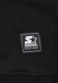 Écusson du logo Starter Black Label en blanc sur tissu tricoté noir avec couture bord côte.