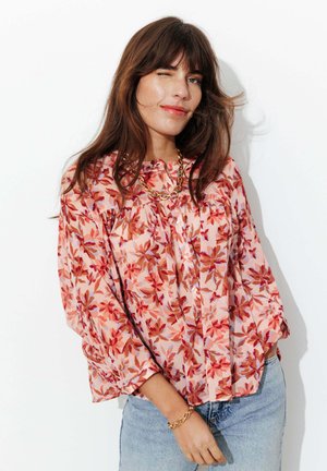 Femme aux longs cheveux bruns portant un chemisier rose à fleurs, un collier en chaîne dorée et un bracelet, ainsi qu'un jean bleu clair, faisant un clin d'œil avec un sourire.