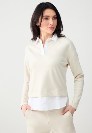 Femme portant un haut à manches longues crème superposé à une chemise blanche à col, avec un pantalon crème assorti, debout les mains dans les poches devant un fond uni.