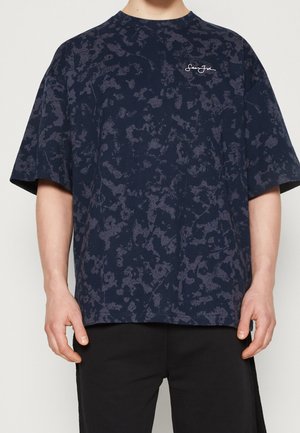 T-shirt oversize bleu marine avec un motif abstrait texturé, col rond, manches courtes et petit logo blanc sur la poitrine.