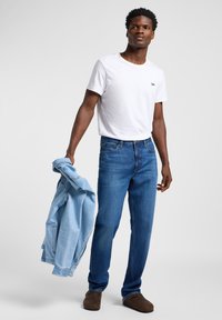 Camiseta blanca de cuello redondo con el logo negro "Lee", jeans de mezclilla azul y una chaqueta de mezclilla azul clara sostenida en una mano. Calzado marrón.