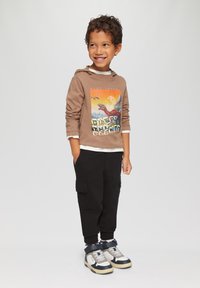 Brauner Hoodie mit Grafikdruck eines Dinosauriers, kombiniert mit schwarzen Cargo-Hosen. Sneaker mit blauen, weißen und beigen Akzenten werden getragen.
