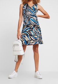 Mekko ilman hihoja, joka on musta ja siinä on värikäs, abstrakti kuvio sinisestä, vaaleanpunaisesta ja keltaisesta. Mukana valkoinen reppu ja valkoiset lenkkarit.