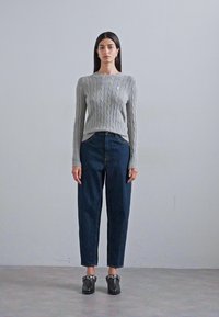 Gray cable-knit sweater, dark blue high-waisted jeans, and black shoes with silver hardware. Model stands against a plain background.