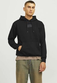 Sudadera negra con un bolsillo tipo canguro en la parte frontal, capucha con cordón y un parche de logo rectangular en el pecho. Llevada sobre una camiseta beige.