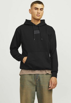 Zwarte hoodie met een voorste kangoeroezak, een capuchon met trekkoord en een rechthoekig logopatch op de borst. Draagt over een beige shirt.