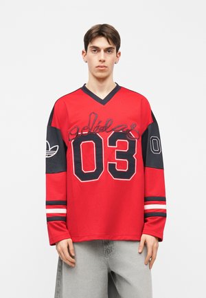 Young man wearing a red and black Adidas long-sleeve sports jersey with "03" and logo details, standing against a plain white background.